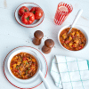 Butternut Squash Ratatouille Soup ON WHITE TABLE WITH SALT & PEPPER, BOWL OF TOMATOES, CHECKERED NAPKIN, SPOONS AND RED CUP.