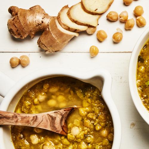 A crock of Channa Potato Stew with a wooden spoon, chopped ginger, and chickpeas