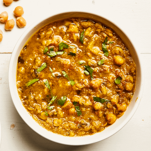 White bowl of Channa Potato Stew Soup with a few chickpeas strewn about on a white background. 