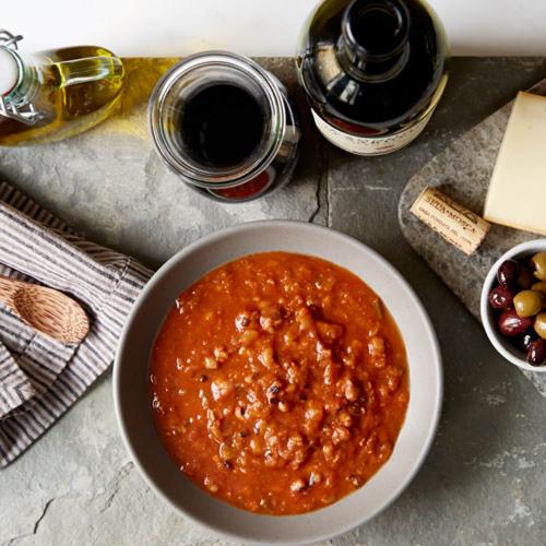 Gray bowl of Black Eyed Pea & Tomato Stew with a striped, linen napkin, wooden spoon, class of wine, bottle of wine, bottle of olive oil, cheese and olives all on a gray background.