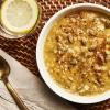 A white bowl of Kabocha Congee soup with a gold spoon, tan napkin, glass of water with lemon, on a woven placemat