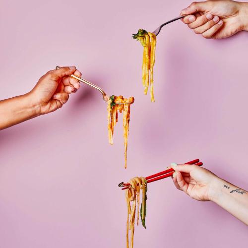 3 hands holding noodles on utensils in front of a purple background.