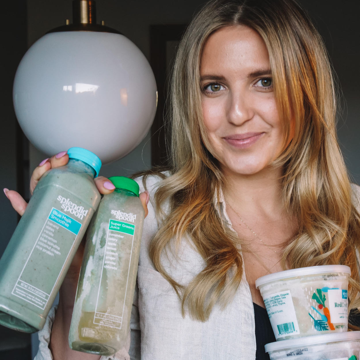 A close-up of a woman looking directly into the camera with a slight grin, holding a variety of Splendid Spoon products in both hands.
