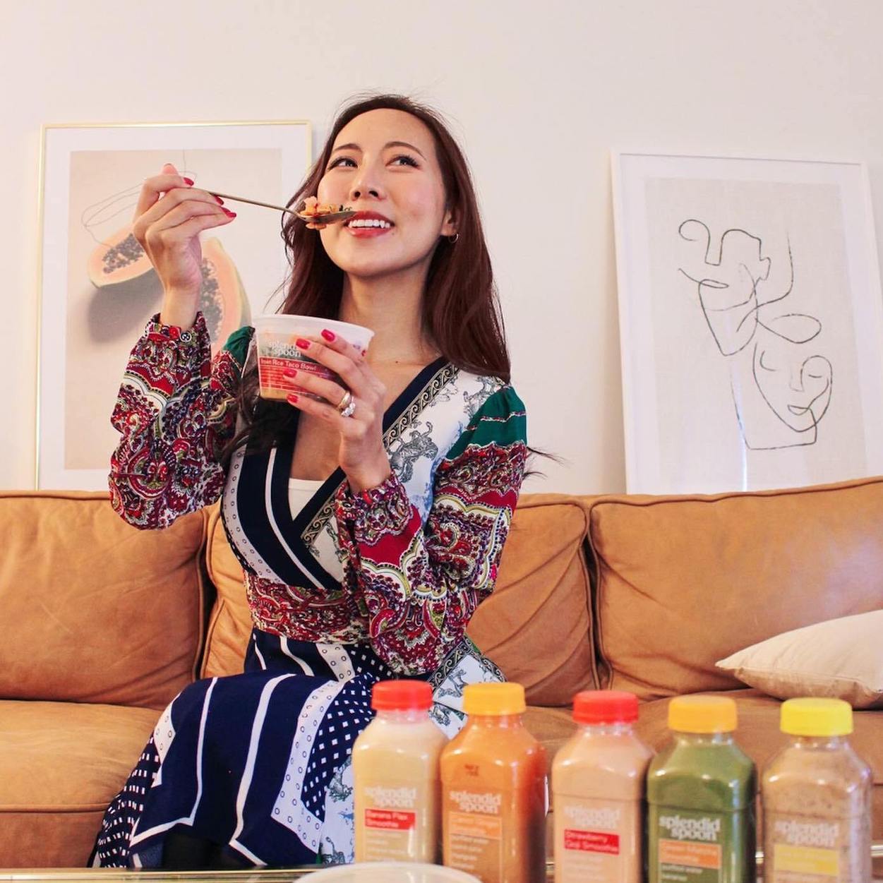 The social media personality J'adore Christina sitting on a brown couch, eating a Splendid Spoon Brown Rice Taco bowl with a spoon. The camera is looking up at her, and she her body is angled slightly to the left, while she looks up and to the right.